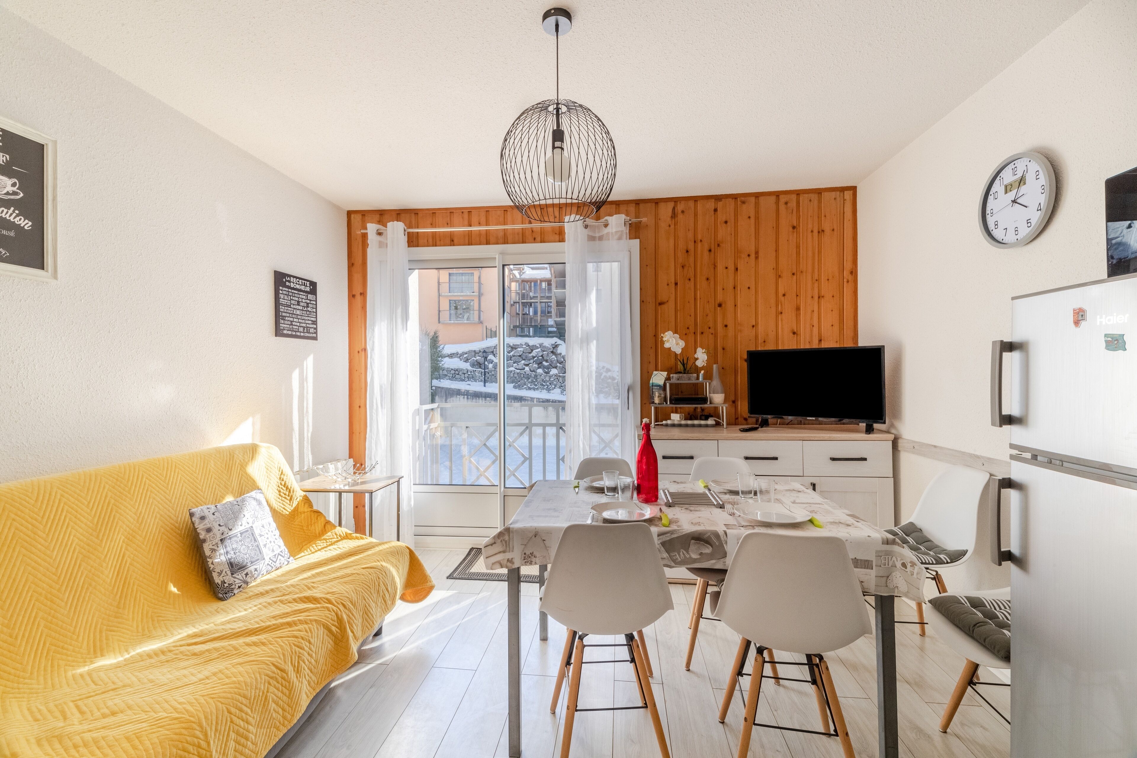 Un salon moderne avec un canapé jaune, une table à manger et des chaises, un téléviseur et un réfrigérateur.