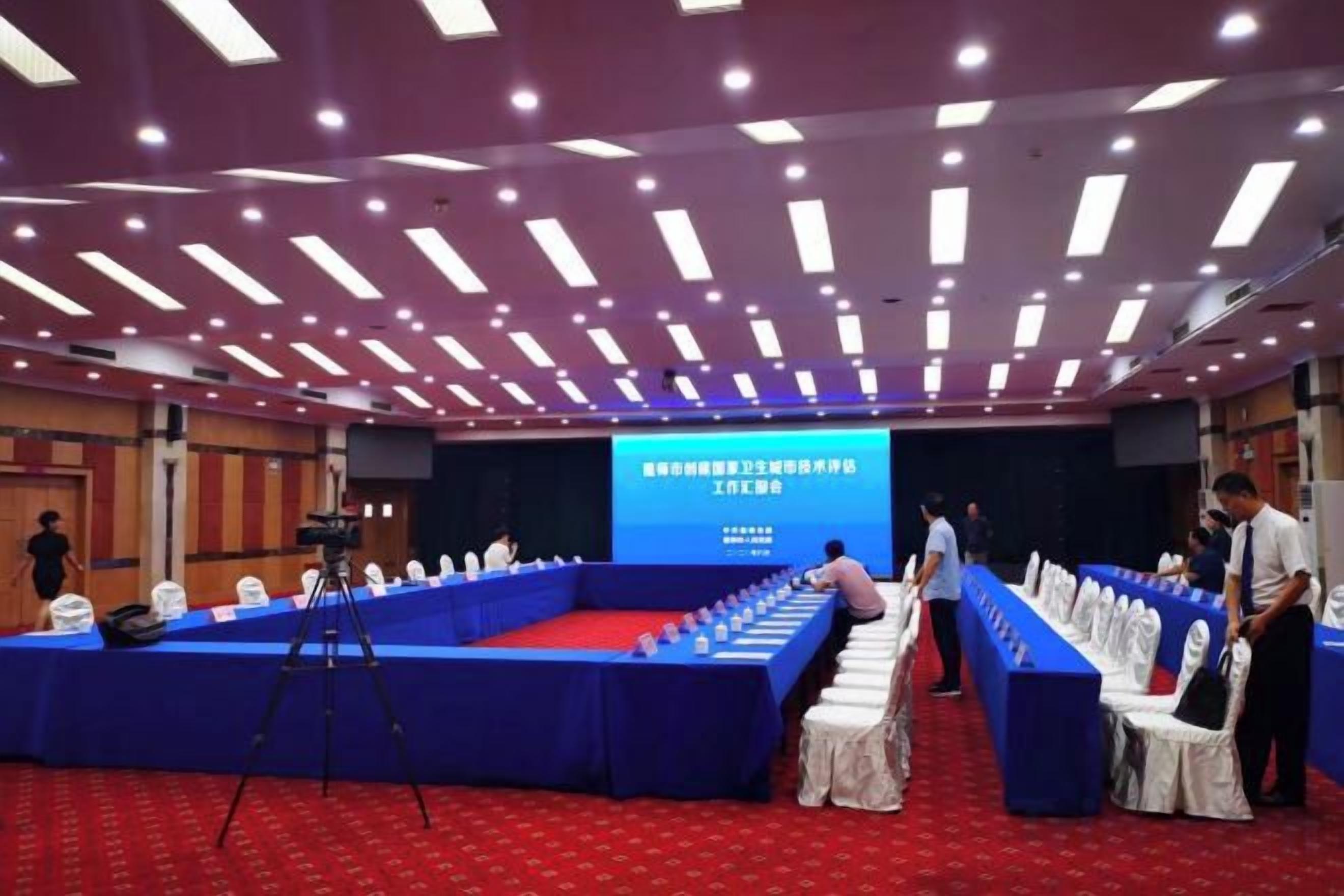 A conference room with a long table, a stage with a screen, and a camera on a tripod.