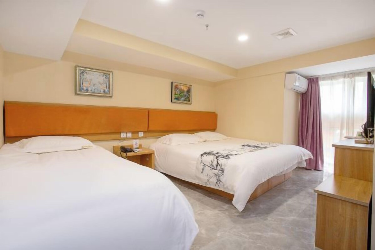 A hotel room with two beds, a wooden headboard, a bedside table with a lamp, and a window with curtains.