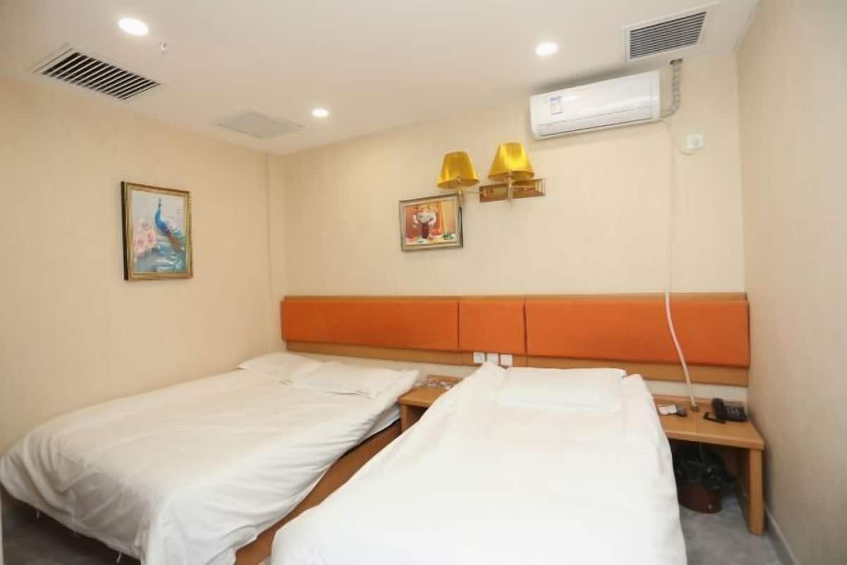 A hotel room with two single beds, a wooden headboard, a bedside table, a wall-mounted air conditioner, and two wall-mounted lamps.