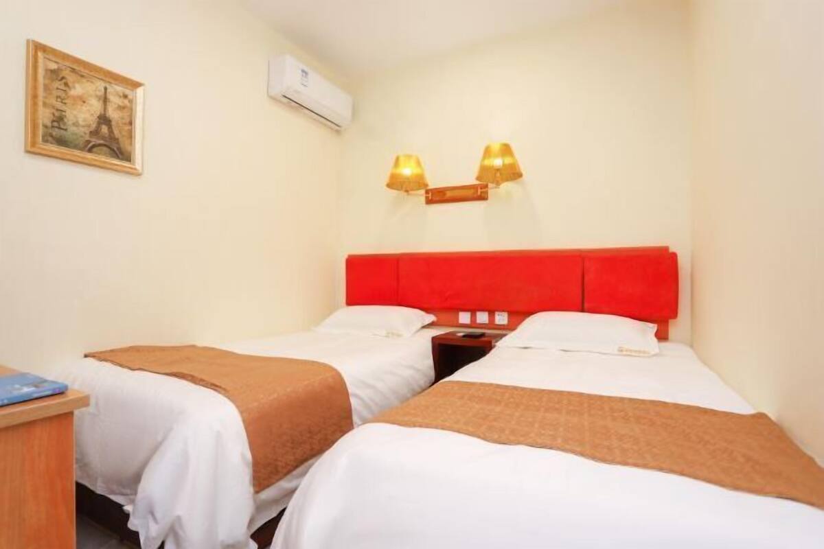 A hotel room with two beds, a desk, a wall-mounted air conditioner, and a painting of the Eiffel Tower.