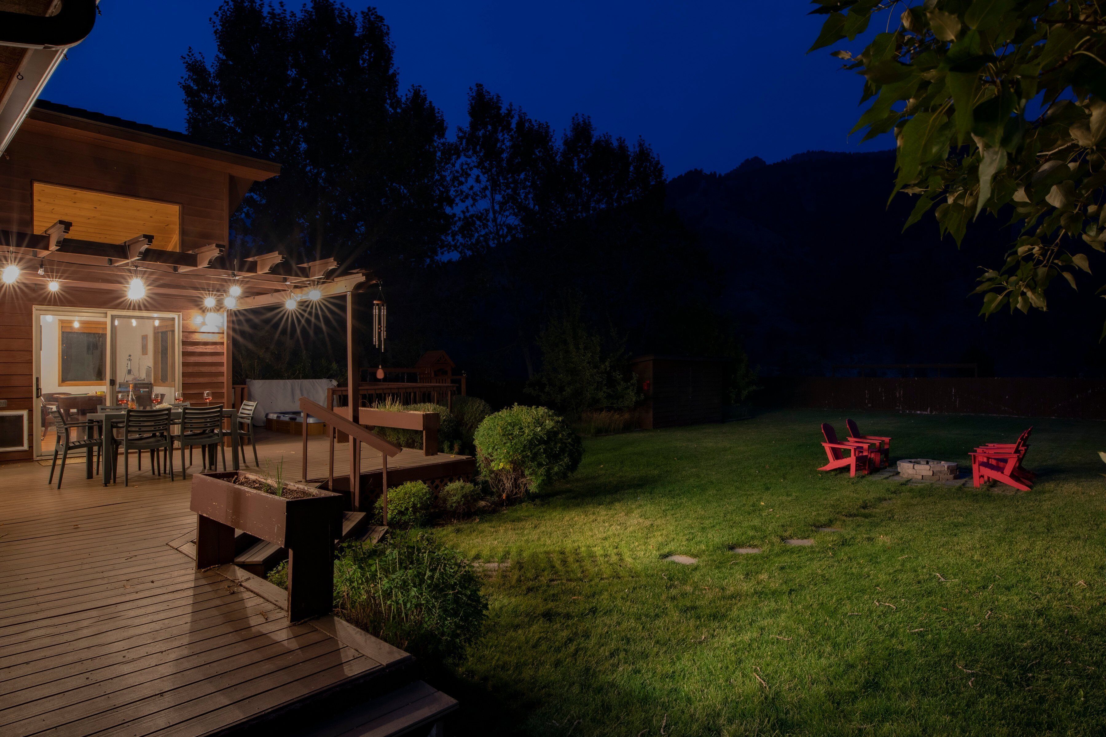 Una terraza de madera con mobiliario exterior y un brasero, rodeada de césped cuidado y árboles.
