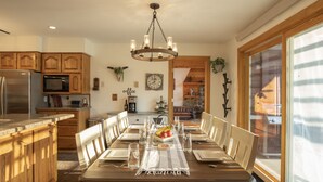 Un coin repas avec une table en bois dressée pour six personnes, entourée de chaises en bois. Un lustre est suspendu au-dessus, et une grande fenêtre laisse entrer la lumière naturelle.
