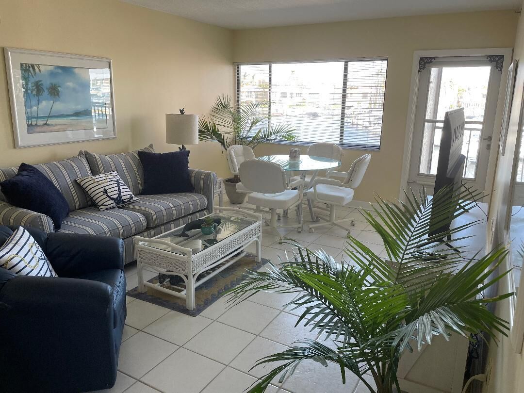 A living room with a striped sofa, a coffee table, and a dining table with chairs. There is a large window with blinds, a door, and a framed picture on the wall.