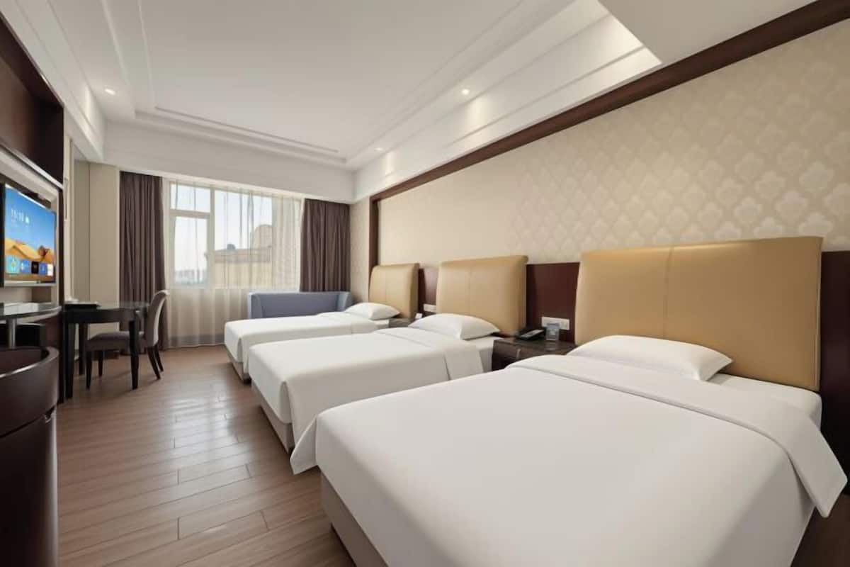 Habitación de hotel con tres camas, un escritorio, un televisor y una ventana con cortinas.