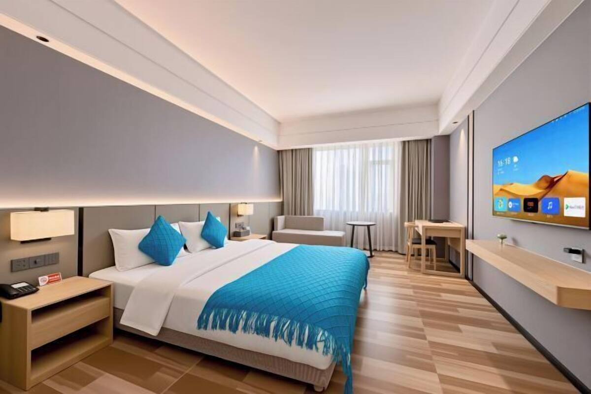 A modern hotel room with a large bed, bedside tables, a desk, a chair, a TV, and a window with curtains.