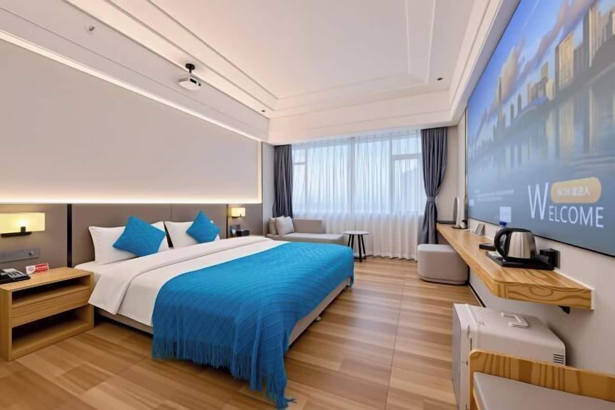 A modern hotel room with a large bed, a TV screen displaying 'WELCOME,' a coffee machine, and a wooden nightstand.