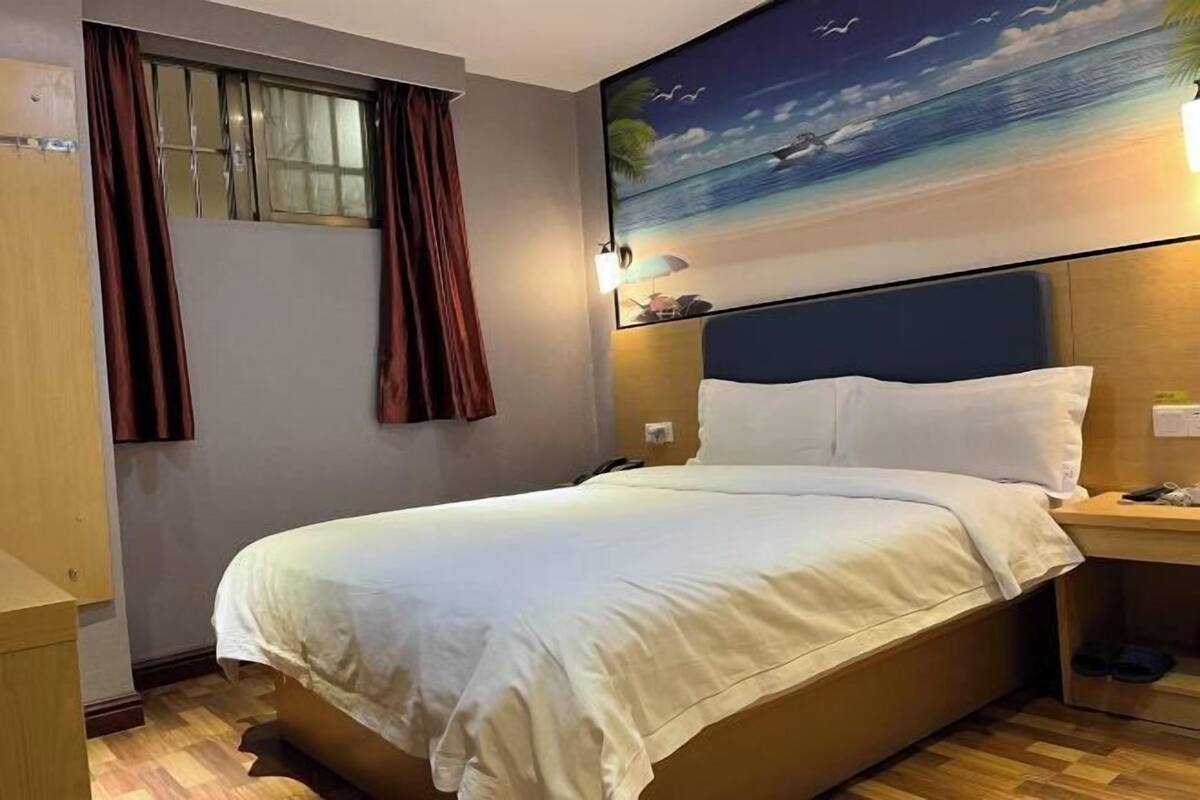 A hotel room with a bed, a nightstand, a window with curtains, and a beach mural on the wall.