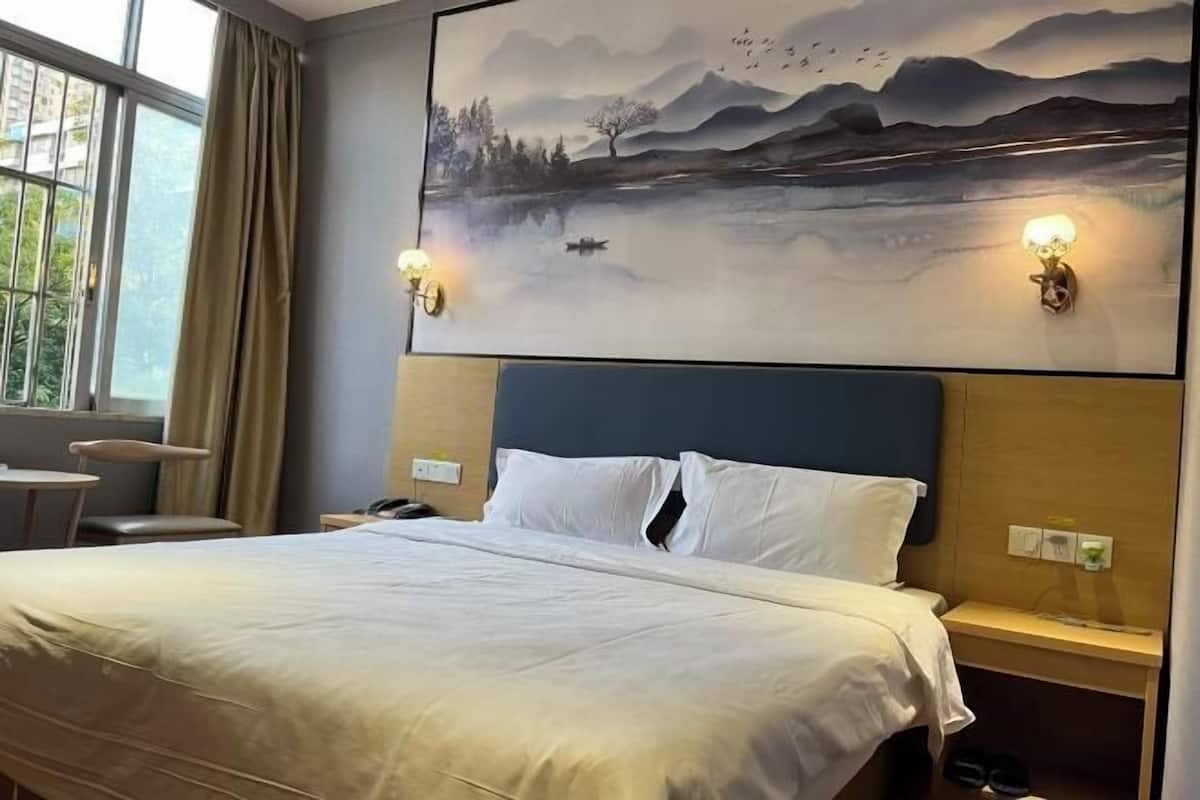 A hotel room with a bed, a desk, a chair, a window with curtains, and a wall-mounted lamp.