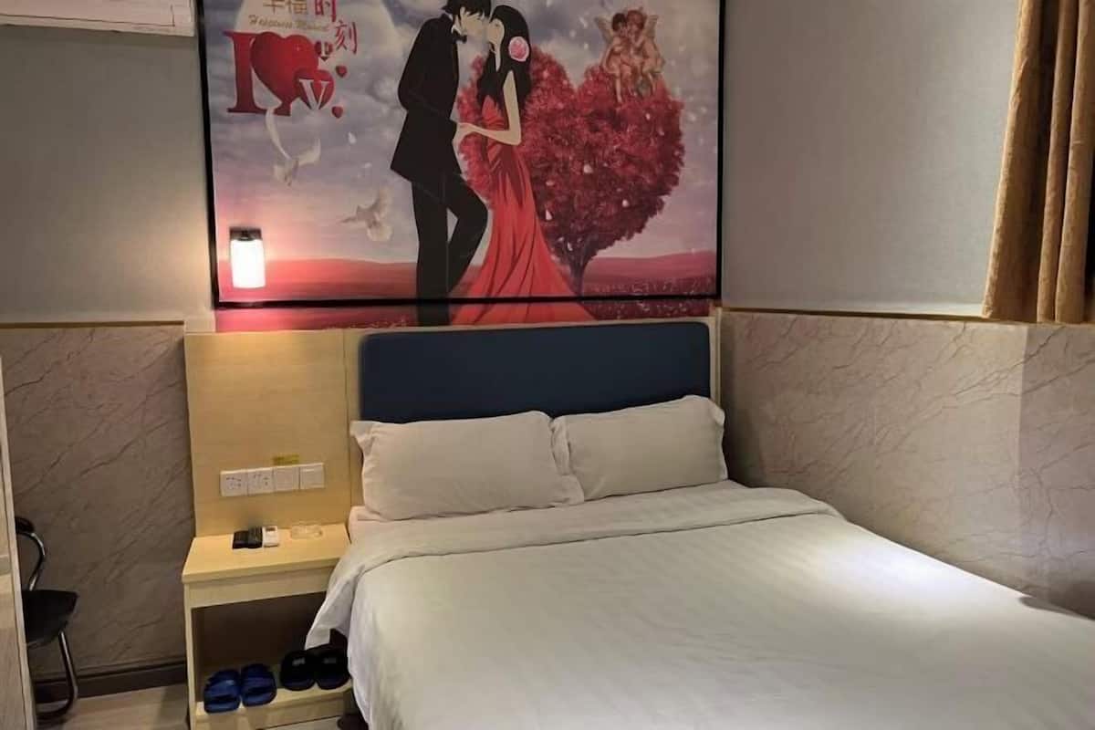 A hotel room with a bed, a nightstand with a lamp, a wall-mounted artwork, and a shoe rack.