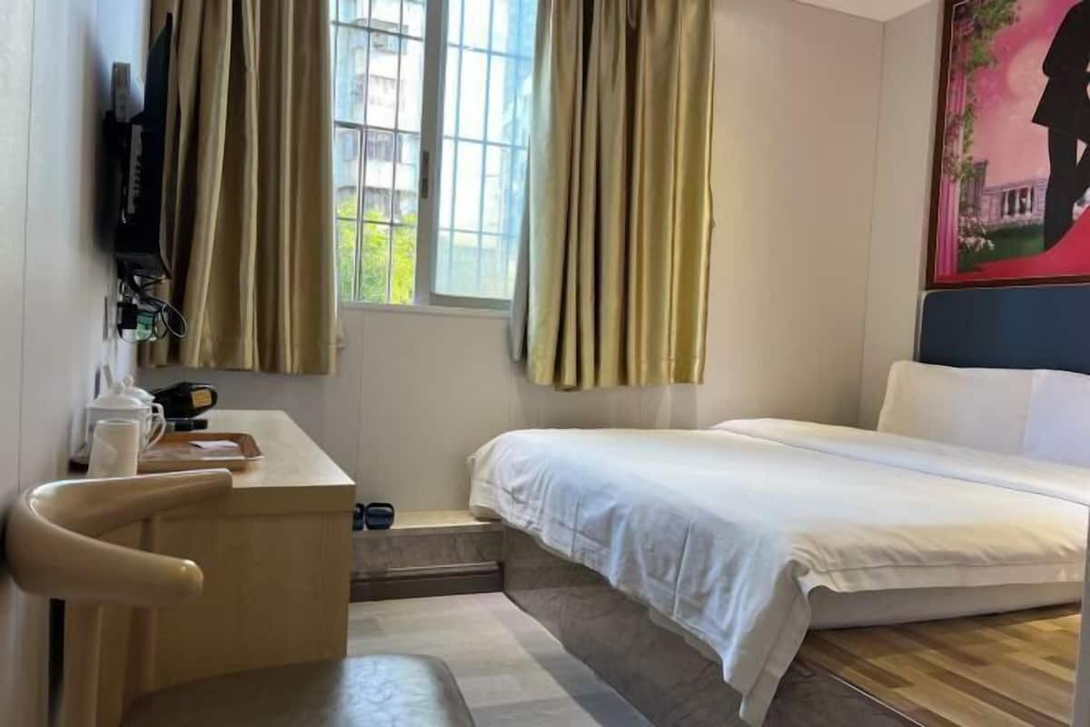 A hotel room with a bed, a desk, a chair, a window with curtains, and a painting on the wall.