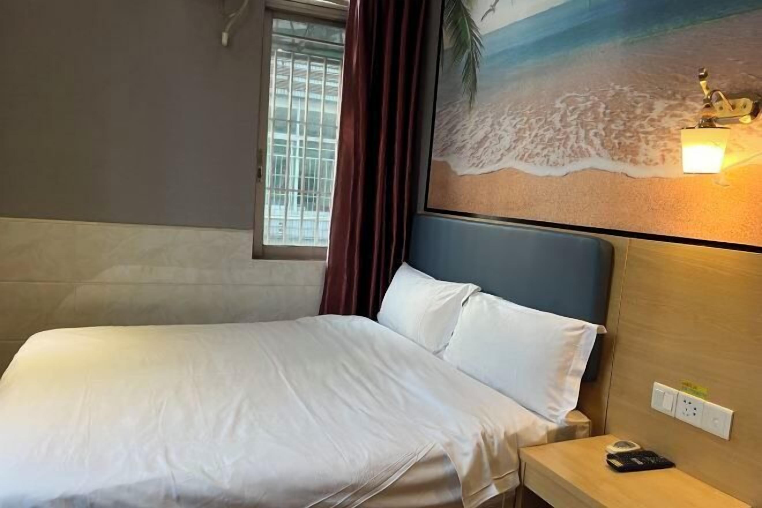 A hotel room with a bed, a nightstand, a wall-mounted lamp, a window with curtains, and a beach-themed painting.