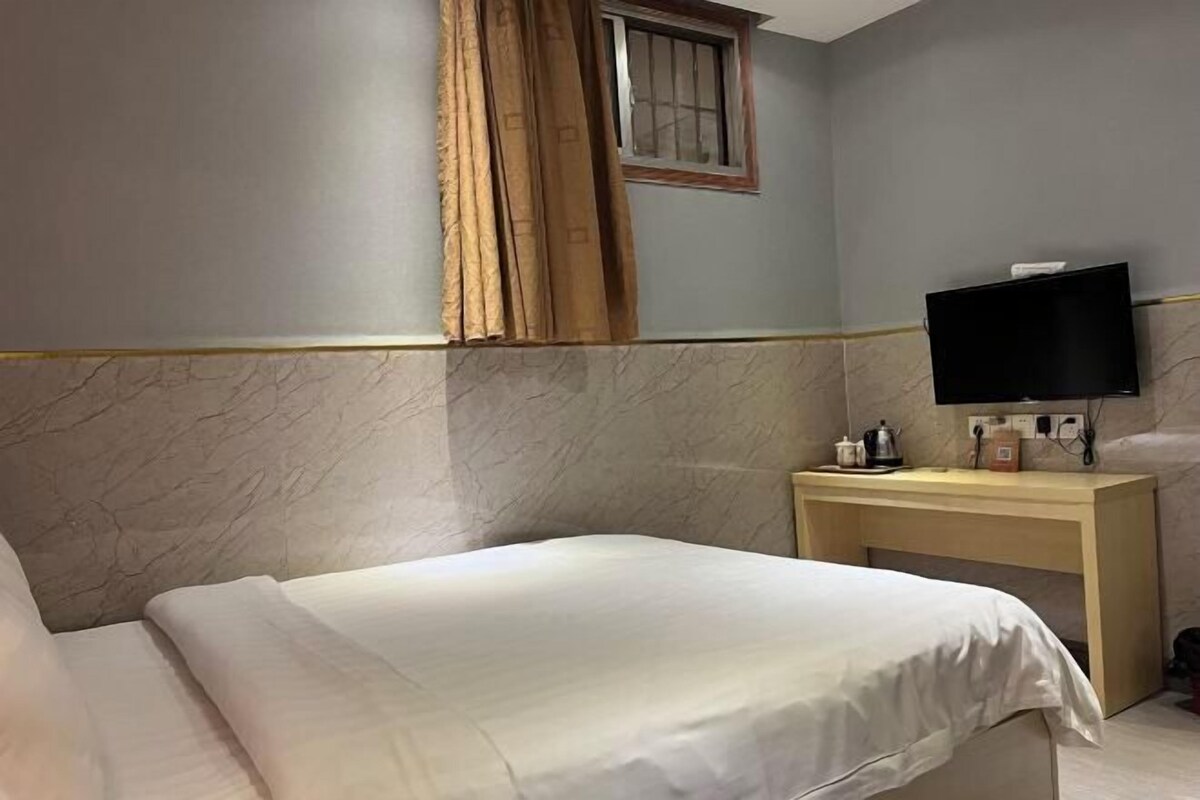 A hotel room with a bed, a TV on a wooden stand, a window with curtains, and a marble wall.
