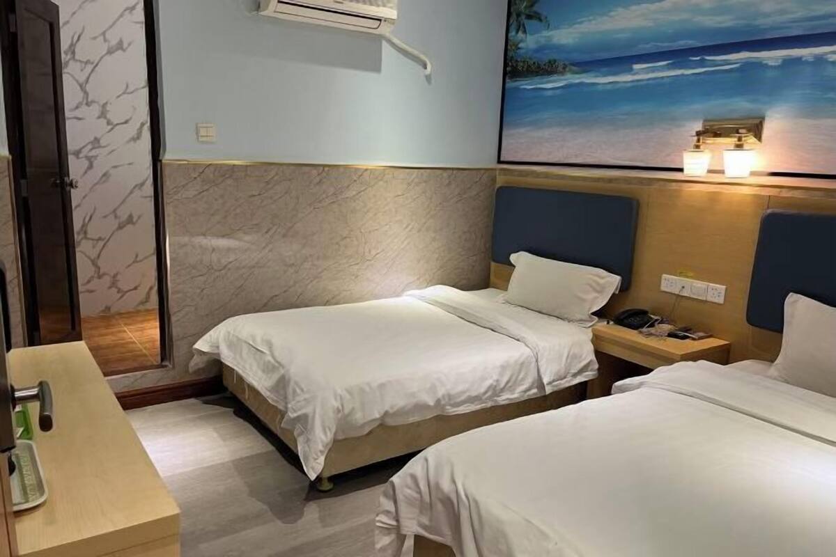 A hotel room with two beds, a desk, a nightstand, an air conditioning unit, and a painting of a beach scene.