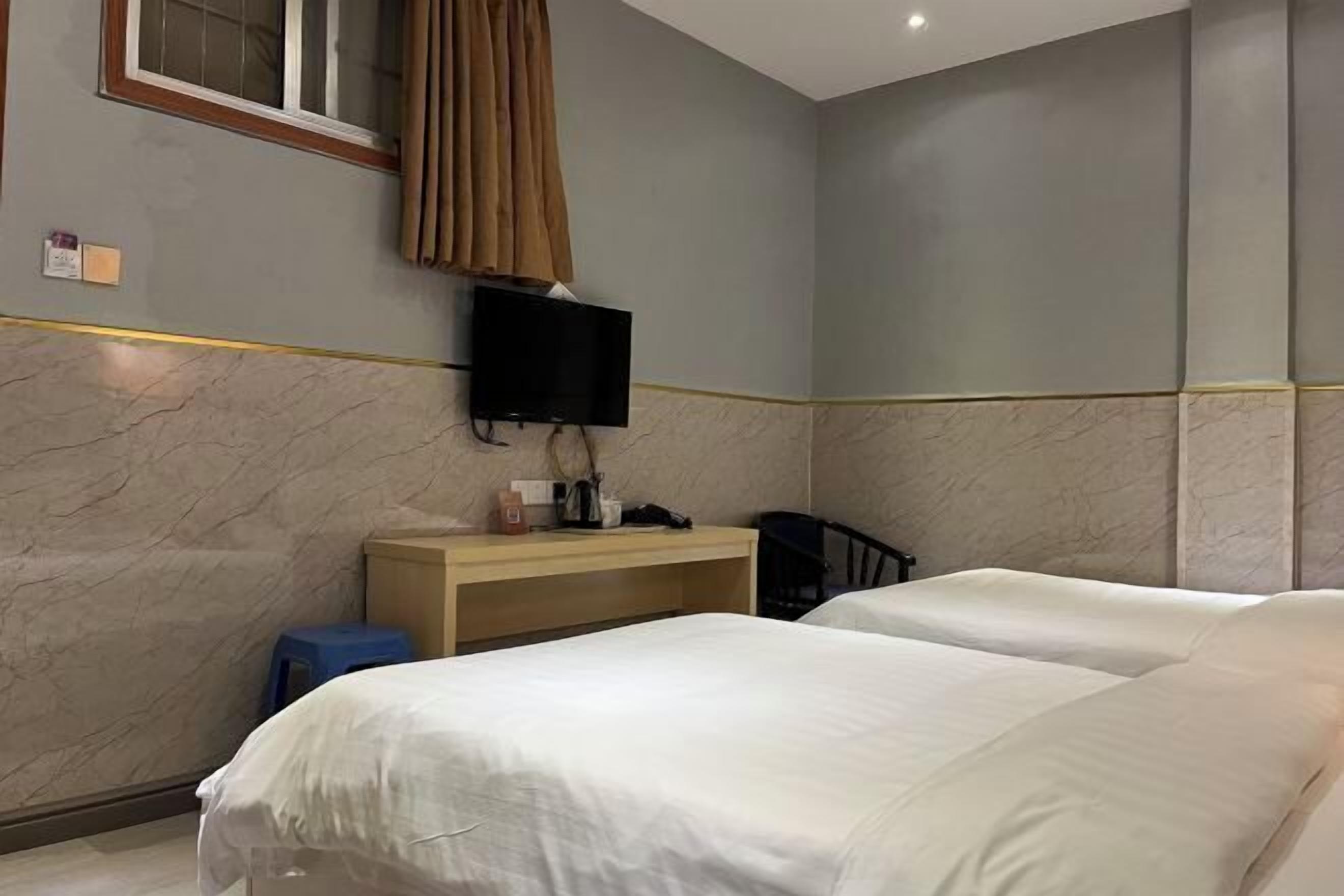 Hotellihuone, jossa on kaksi sänkyä, seinälle asennettu televisio, puinen työpöytä ja pieni sininen jakkara.