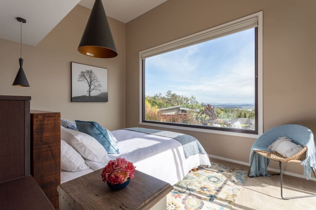 A bedroom with a large window, a bed with white and blue bedding, a blue chair, and a wooden nightstand with a vase of flowers.