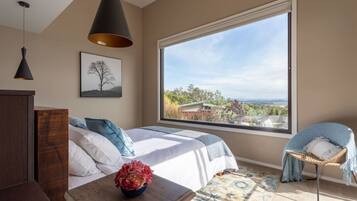 A bedroom with a large window, a bed with white and blue bedding, a blue chair, and a wooden nightstand with a vase of flowers.