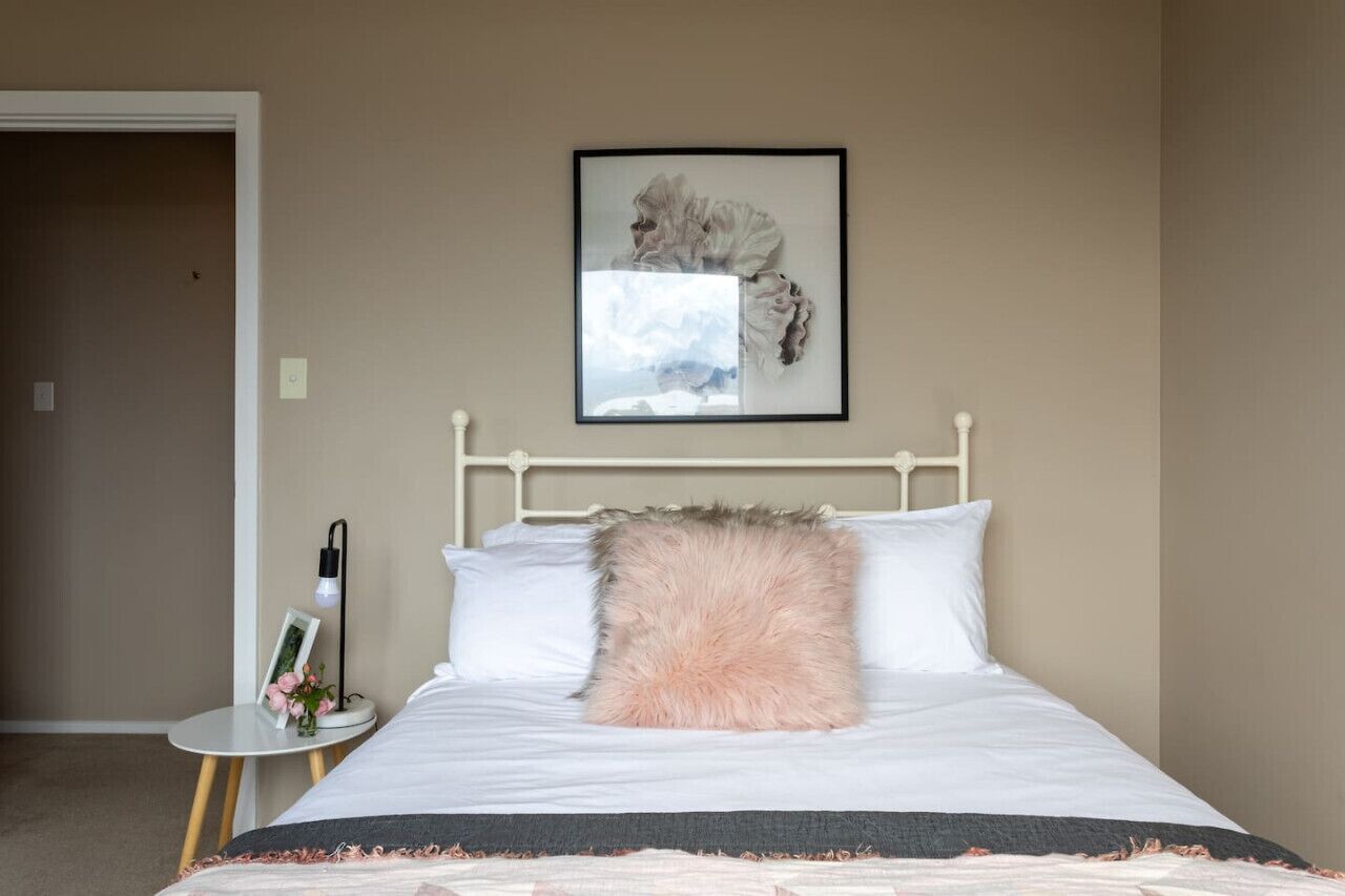 Un lit bien fait, avec des draps blancs et un oreiller rose moelleux, une table de chevet avec une lampe et un cadre photo, et une œuvre d’art encadrée accrochée au mur au-dessus du lit.
