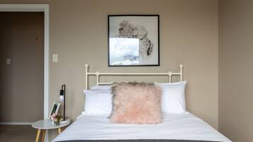 Un lit bien fait, avec des draps blancs et un oreiller rose moelleux, une table de chevet avec une lampe et un cadre photo, et une œuvre d’art encadrée accrochée au mur au-dessus du lit.
