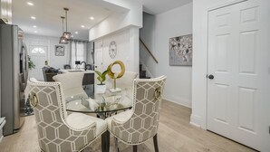 A modern dining area with a glass table and patterned chairs, a kitchen in the background, and a white door. - Modern Niagara-area Spa-like Home (St. Catharines)