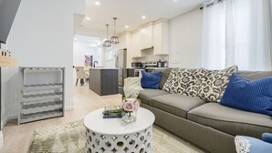 A modern living room with a grey sofa, patterned cushions, a white coffee table with books and flowers, and a kitchen in the background. - Modern Niagara-area Spa-like Home (St. Catharines)
