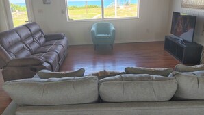 A living room with a brown leather sofa, a light blue armchair, and a television on a stand. A large window offers a view of the beach. - Melbourne Beach House and Basement Apt on 1.2 acres ocean front (Melbourne Beach)