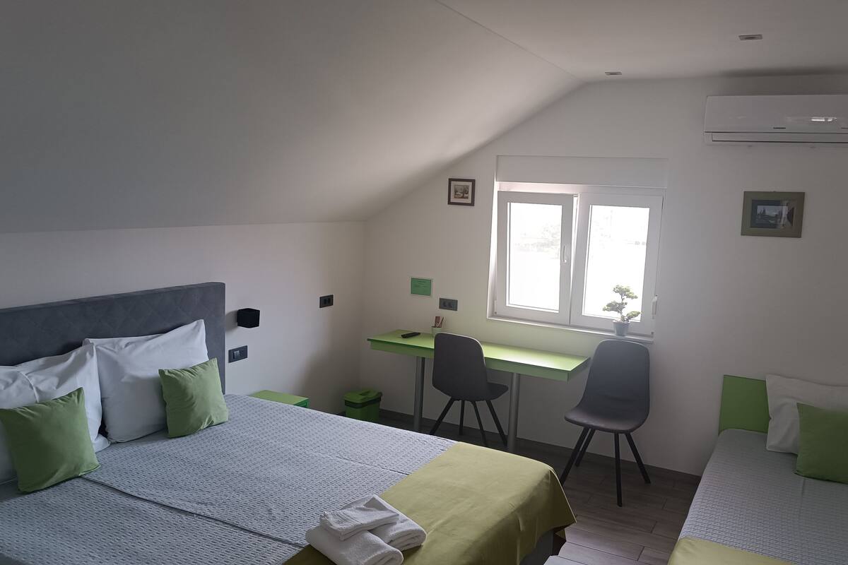 Un dormitorio con cama, escritorio y silla. Se ve una ventana con vistas a árboles y un edificio.