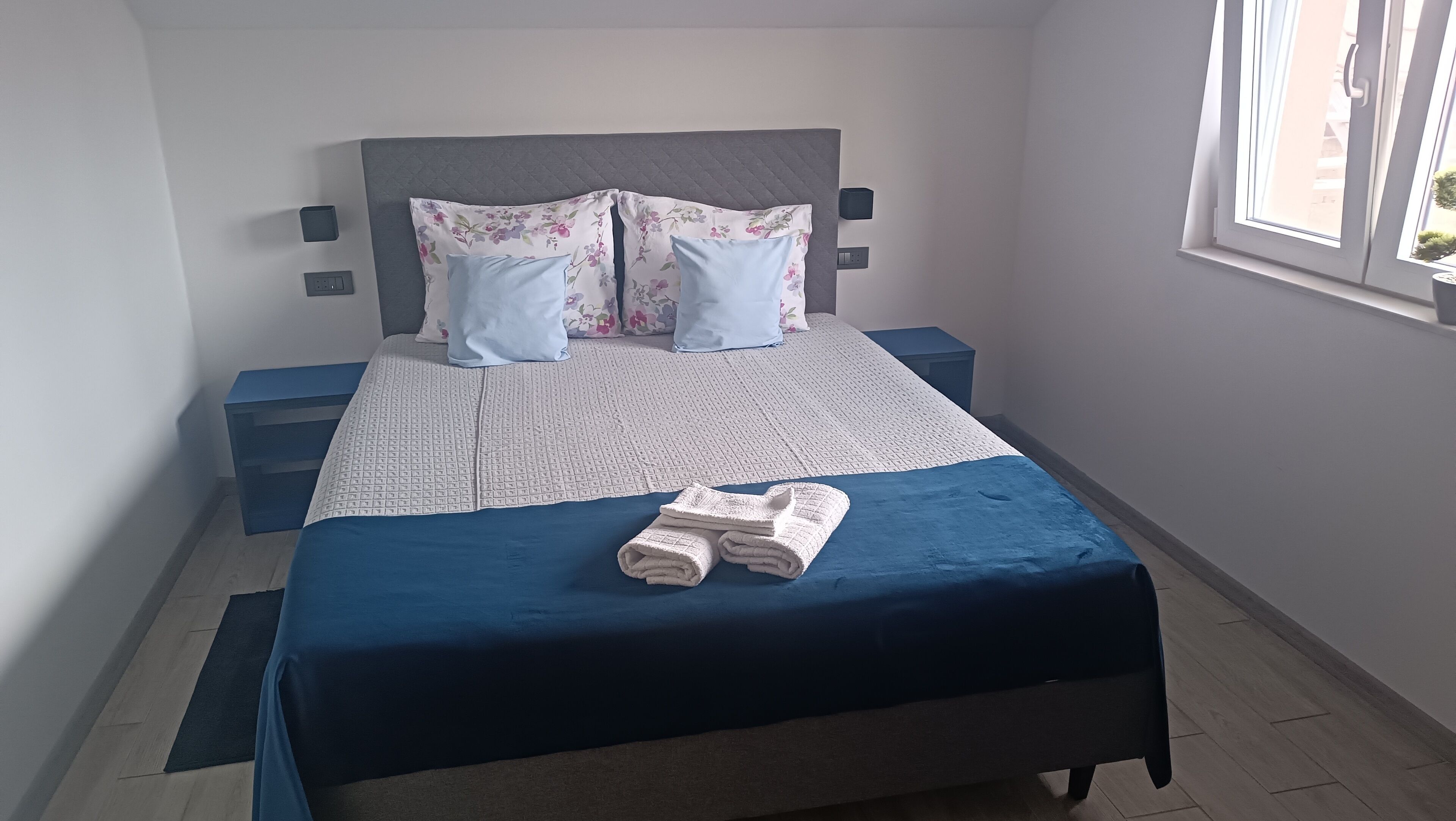 Una cama bien hecha con una colcha azul, una toalla doblada, un cabecero gris y almohadas estampadas con motivos florales.