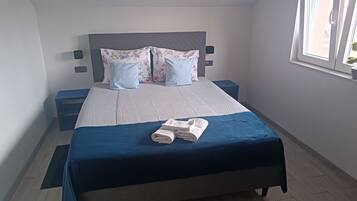 Una cama bien hecha con una colcha azul, una toalla doblada, un cabecero gris y almohadas estampadas con motivos florales.