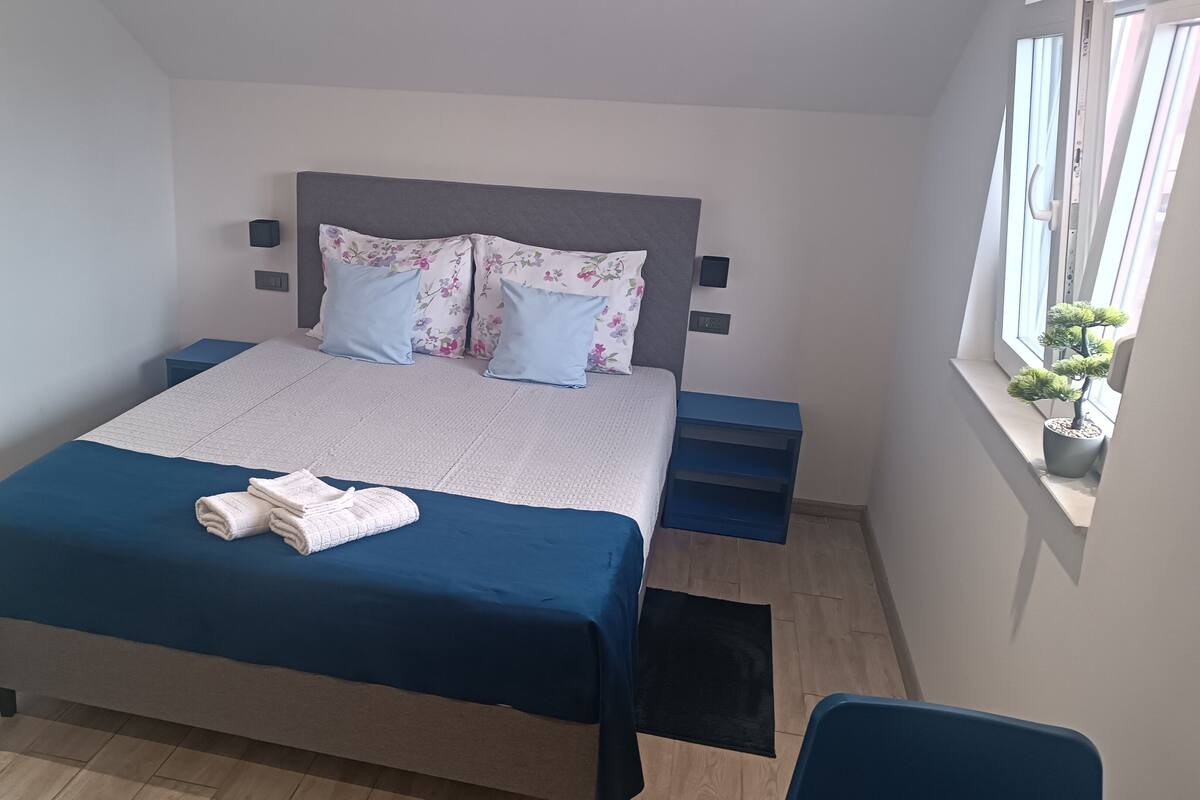 Una cama bien hecha con una colcha azul y almohadas blancas, una mesita de noche, una ventana con una planta en maceta y una silla.
