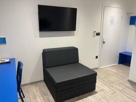 Una habitación con una televisión de pantalla plana montada en la pared, un sofá negro, un escritorio azul con una silla y una puerta con cerradura de llave electrónica.