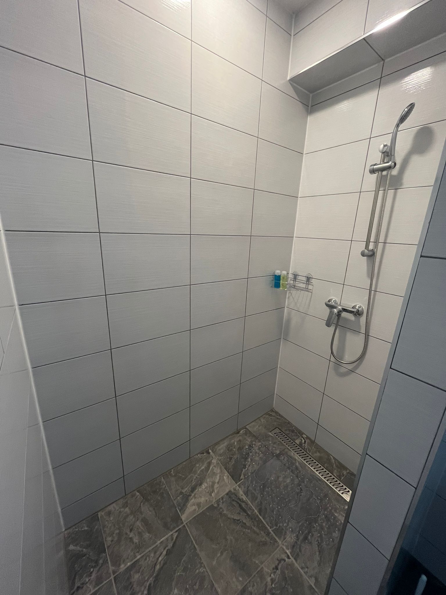 Un petit espace de douche carrelé, équipé d’un pommeau de douche à main et d’un robinet fixé au mur.