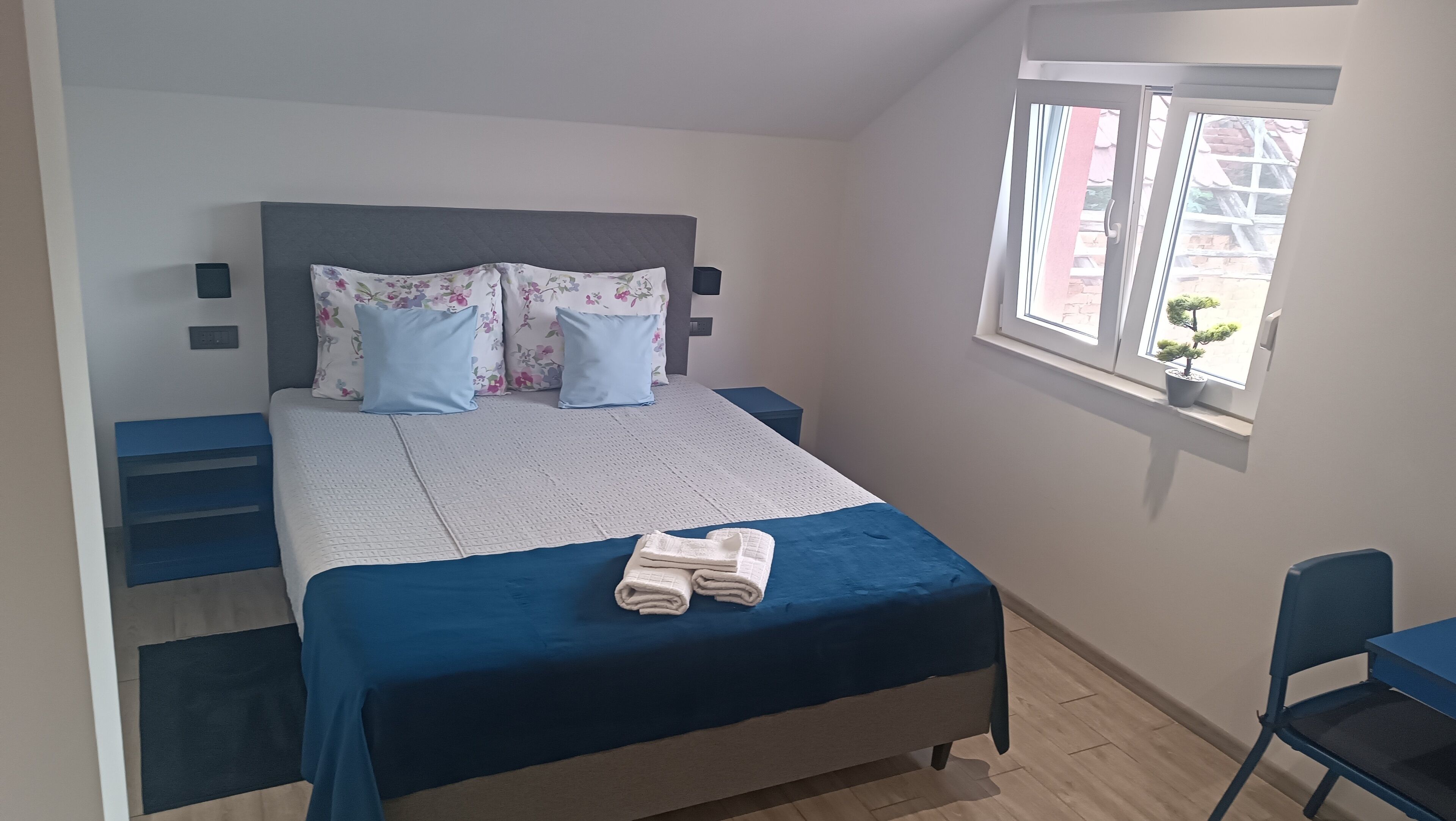 Una cama bien hecha con ropa de cama azul, una ventana con vista a edificios y un escritorio pequeño con una silla.