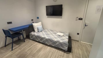 A small, single-bed room with a blue desk, a wall-mounted TV, and a clock.