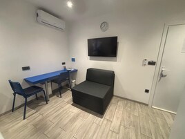 Una habitación pequeña con un escritorio azul, dos sillas, un sofá negro, un televisor montado y una puerta.