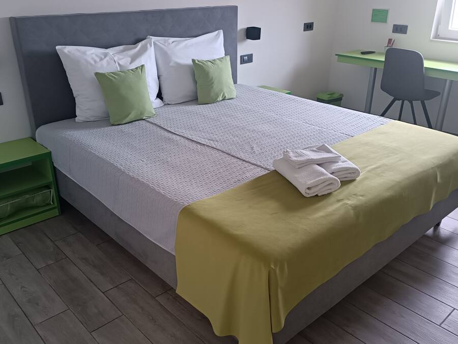 Un lit bien fait, recouvert d’une couverture verte et agrémenté de serviettes blanches, une table de chevet verte, un bureau avec une chaise et une fenêtre.