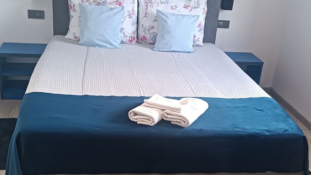 Una cama bien hecha con una colcha azul, dos almohadas con motivos florales y una pila de toallas dobladas sobre la cama.