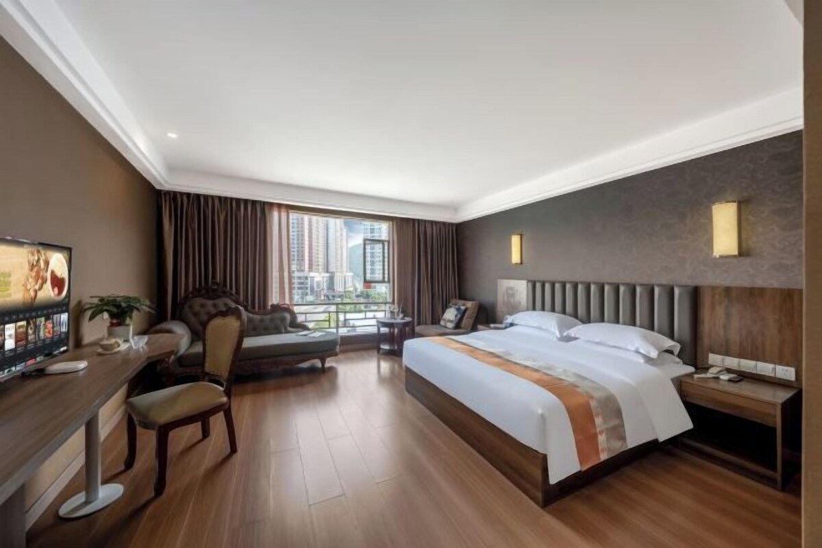 A hotel room with a large bed, a desk with a computer, a chair, a TV, and a window with a city view.