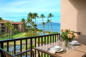 A balcony with a table set for dining, overlooking a pool and a golf course with palm trees, and a view of the ocean.