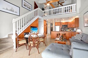 A spacious living area with a dining table set for four, a grey sofa, and a kitchen with wooden cabinets and a central island.