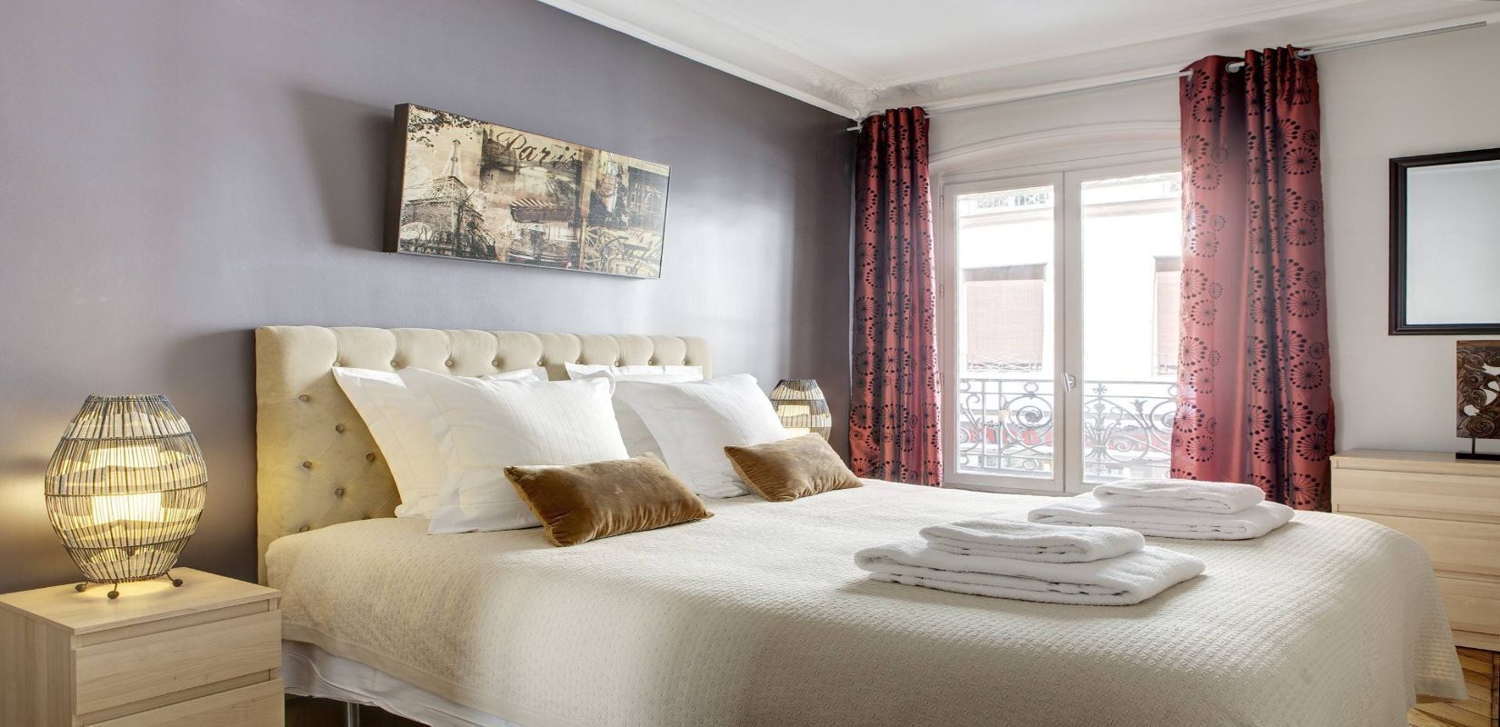 A neatly made bed with white linens and a beige headboard, a bedside lamp, and a folded towel on the nightstand.