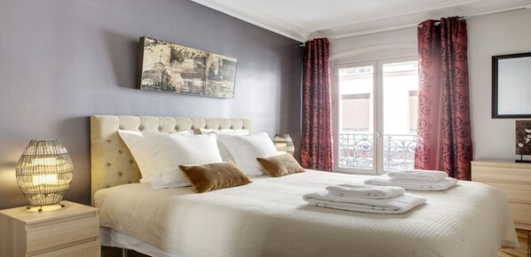 A neatly made bed with white linens and a beige headboard, a bedside lamp, and a folded towel on the nightstand.