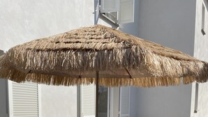 Uma mesa de piquenique de madeira com um toldo de palha, cercada por um muro branco e uma janela com portadas.