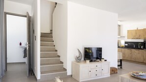 Uma sala de estar moderna com escada, uma TV de tela plana em um console branco e uma área de cozinha com armários de madeira.