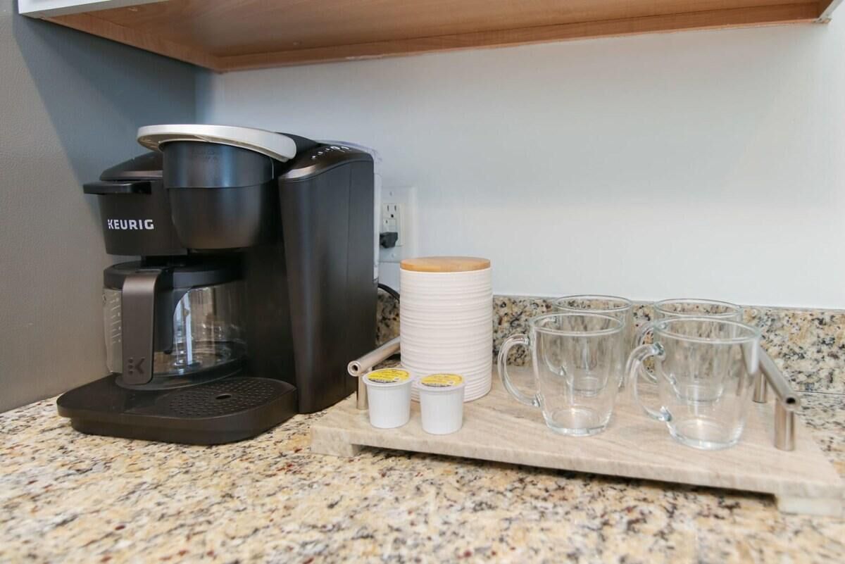 A Keurig coffee machine on a granite countertop with four clear mugs and two small containers, likely for coffee pods, on a wooden tray.
