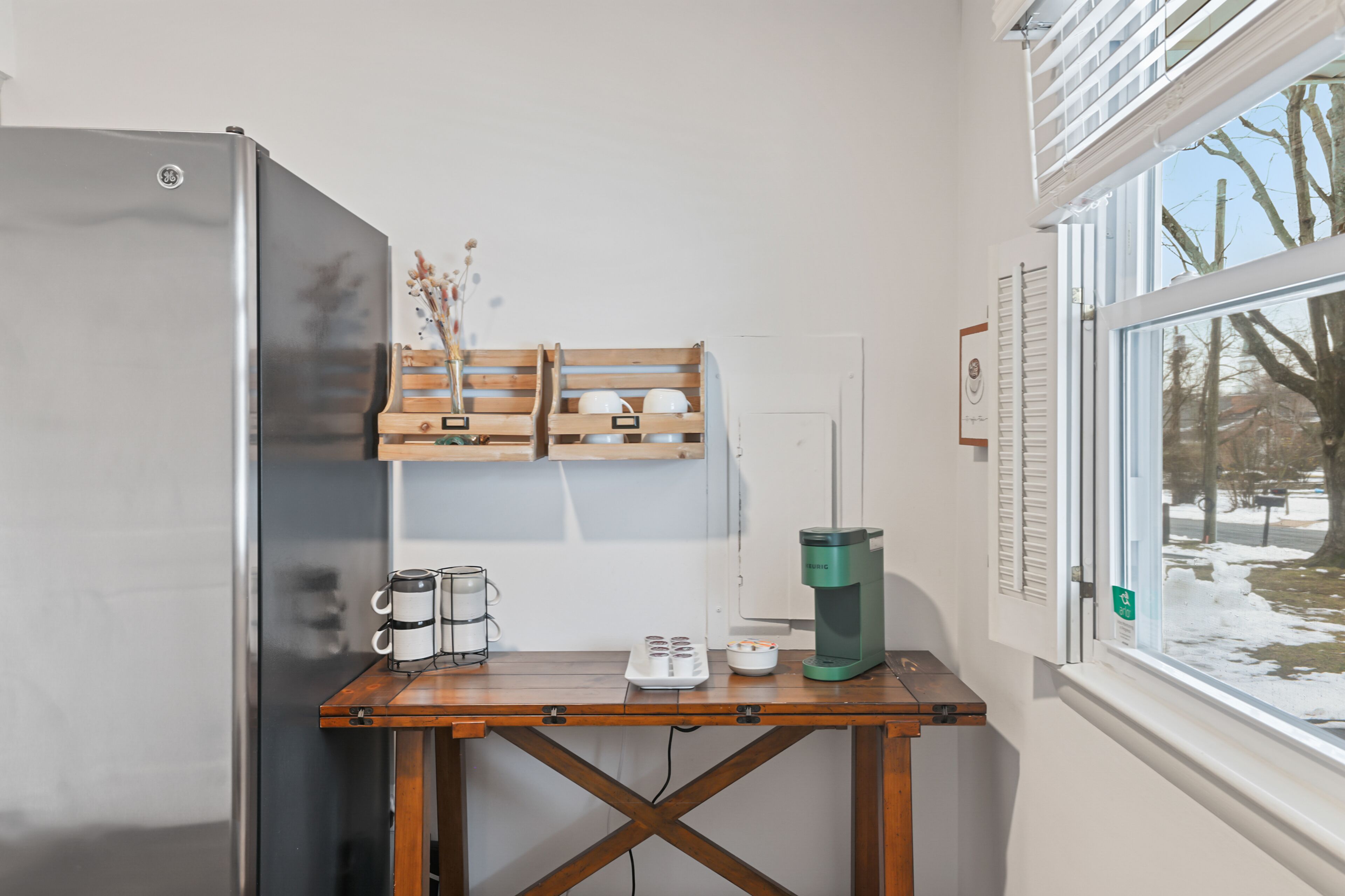 Une petite cuisine équipée d’un réfrigérateur en acier inoxydable, d’une table en bois avec une machine à café, et d’une fenêtre donnant sur des arbres et de la neige.