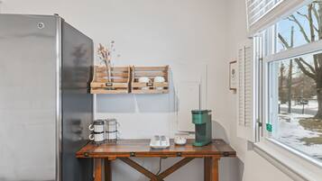 Une petite cuisine équipée d’un réfrigérateur en acier inoxydable, d’une table en bois avec une machine à café, et d’une fenêtre donnant sur des arbres et de la neige.