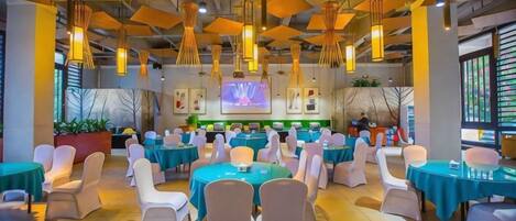 Une salle de conférence avec des tables et des chaises rondes, une scène avec un écran et une grande fenêtre donnant sur des plantes.