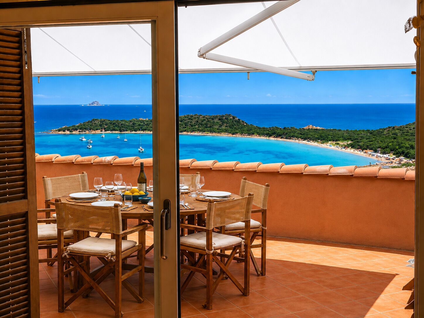 Un balcon ouvert avec une table dressée pour le dîner, offrant une vue sur un paysage côtier avec une eau d’un bleu limpide et des bateaux.