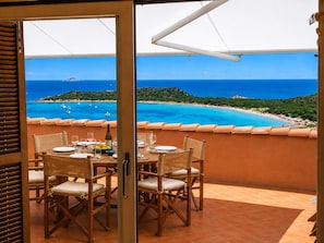 An open balcony with a table set for dining, overlooking a coastal landscape with clear blue waters and boats.